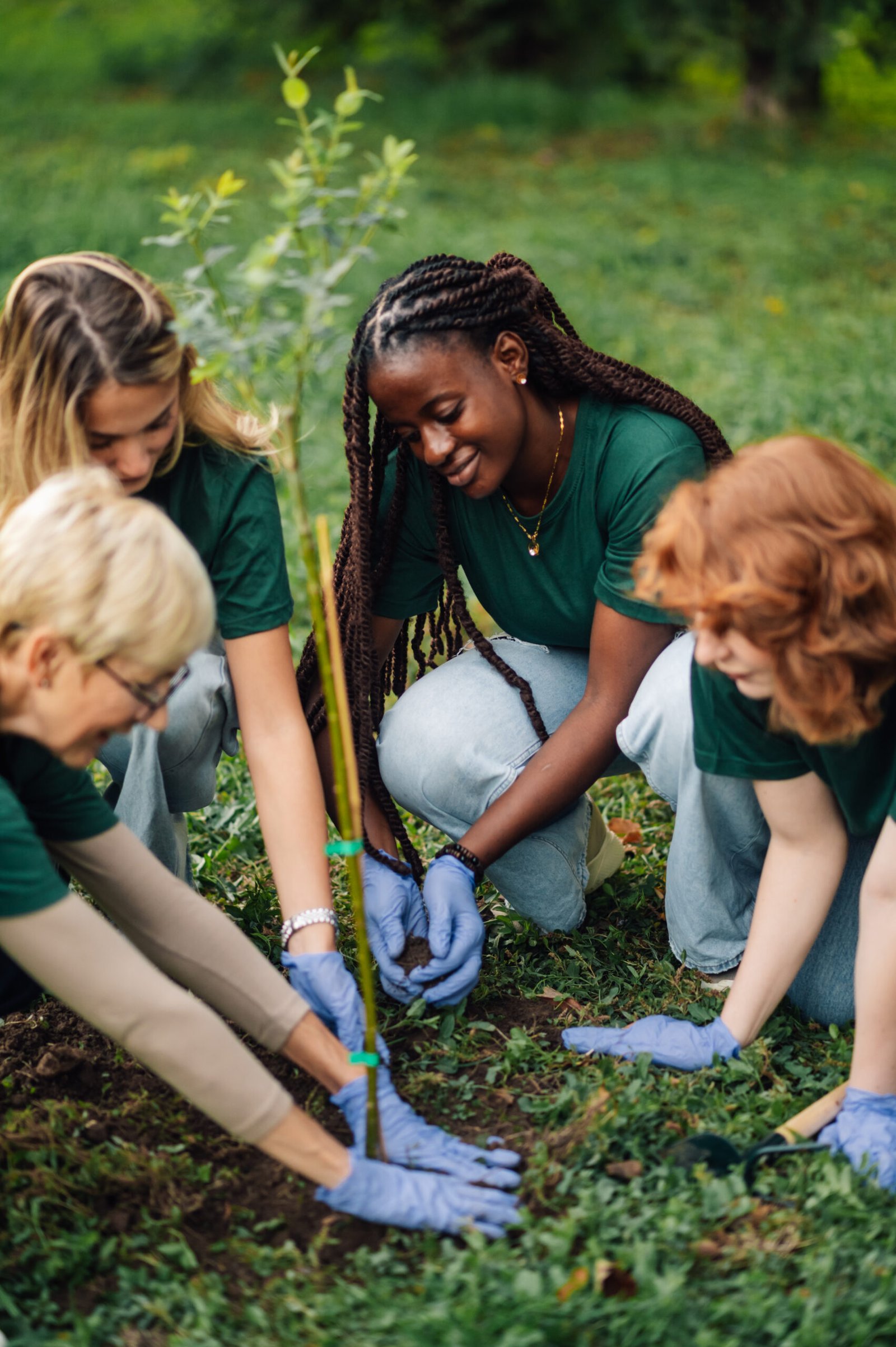 Diverse group of volunteers wearing green shirts is planting a tree in a park, working together to protect the environment